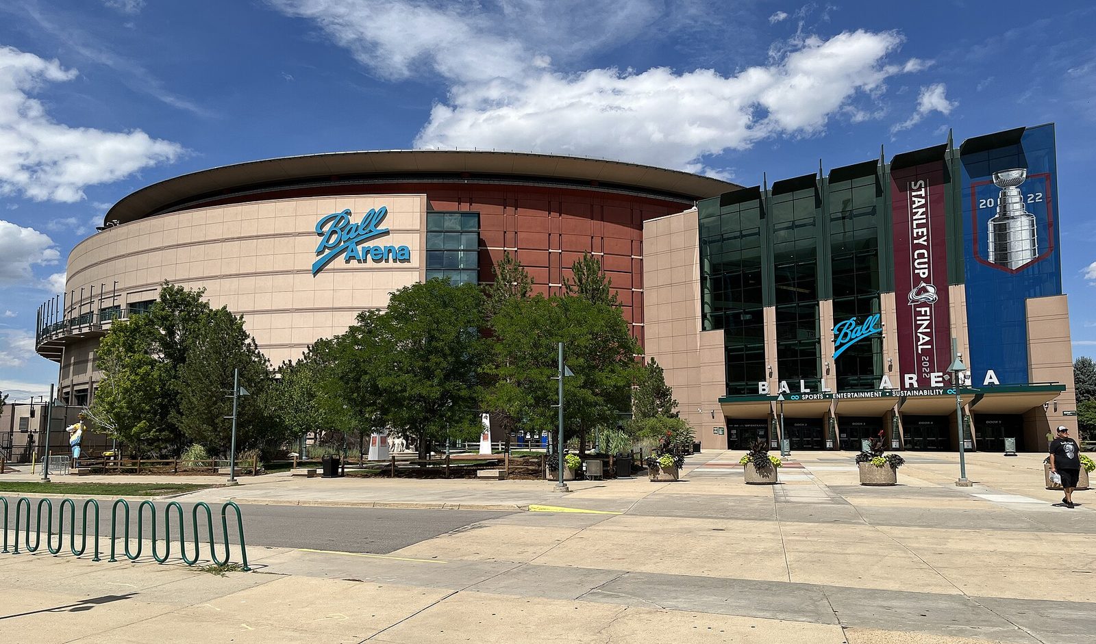 The exterior of Ball Arena in Denver on a summer day, with the arena's curved beige and red facade, a Stanley Cup Final banner on the glass entrance tower and bike racks in the foreground