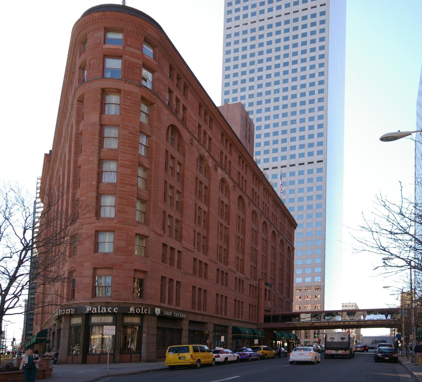 The triangular red sandstone exterior of the Brown Palace Hotel in Denver on its corner lot, with the Ship Tavern sign at street level and a modern glass tower behind