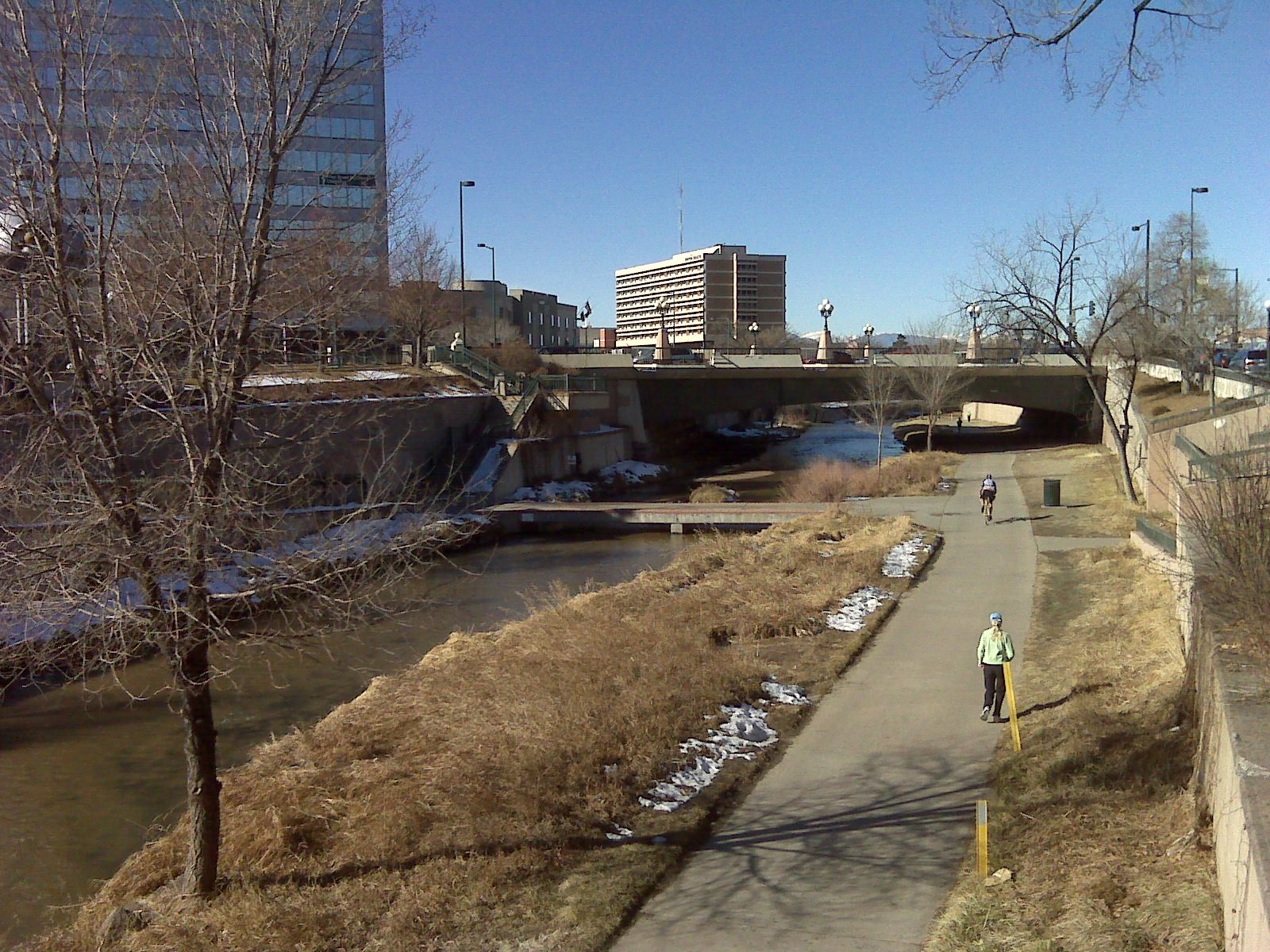 The paved Cherry Creek trail running below Speer Boulevard in Denver on a clear winter day, with a cyclist and a walker on the path, patches of old snow on the banks and downtown buildings behind