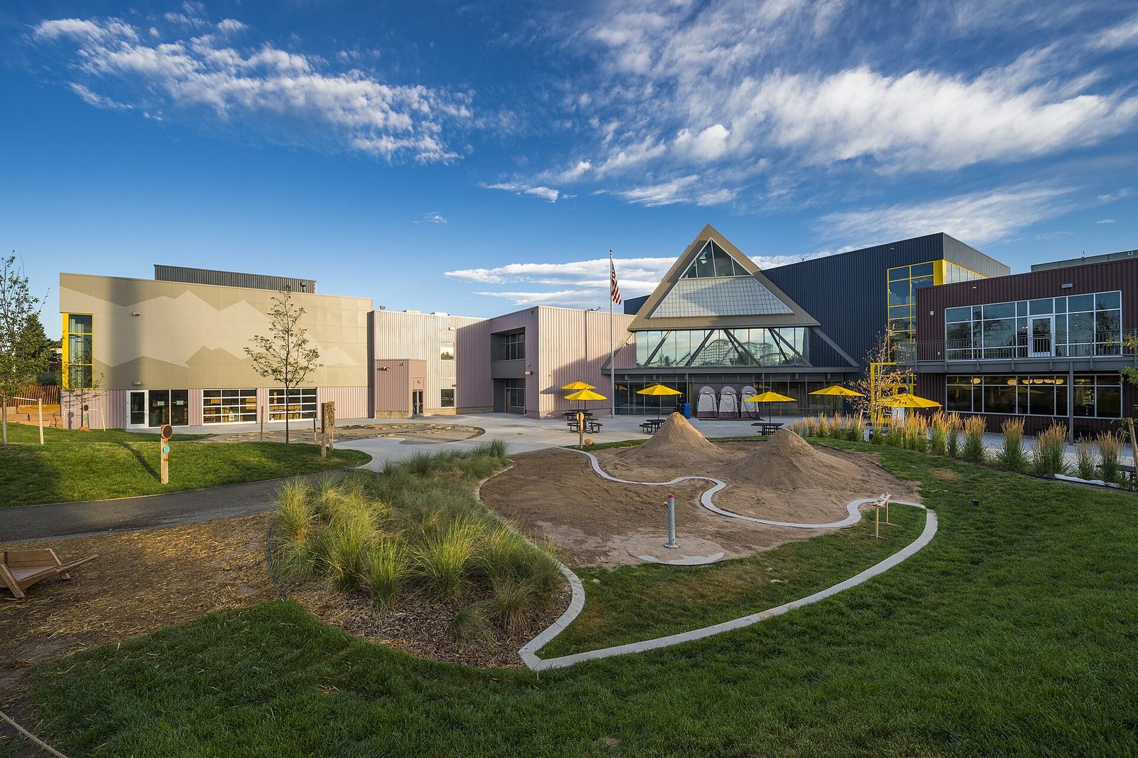 The Children's Museum of Denver at Marsico Campus seen across its outdoor play area, with sand mounds shaped like mountains, a water-play channel, yellow umbrellas and the angular museum buildings behind