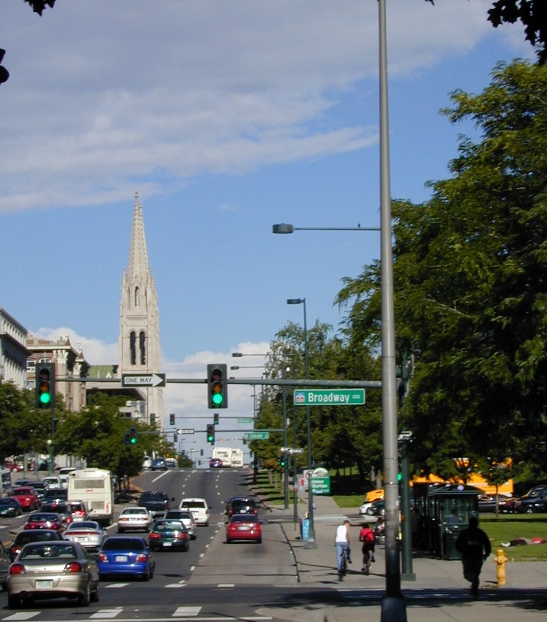 Colfax Avenue in Denver looking east toward the intersection with Broadway, with a Broadway street sign over the road, traffic in both directions, cyclists on the right and the stone spire of a cathedral in the distance
