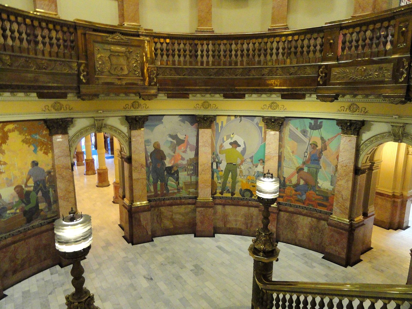 The interior rotunda of the Colorado State Capitol seen from an upper level, with carved wooden balustrades, rose onyx columns and a series of painted murals showing workers, machinery and construction scenes