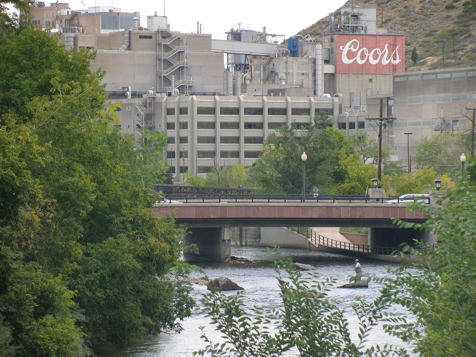 The industrial buildings of the Coors brewery in Golden, Colorado, with a large Coors sign on a red tank, rising above Clear Creek where a fly fisher stands in the water below a road bridge