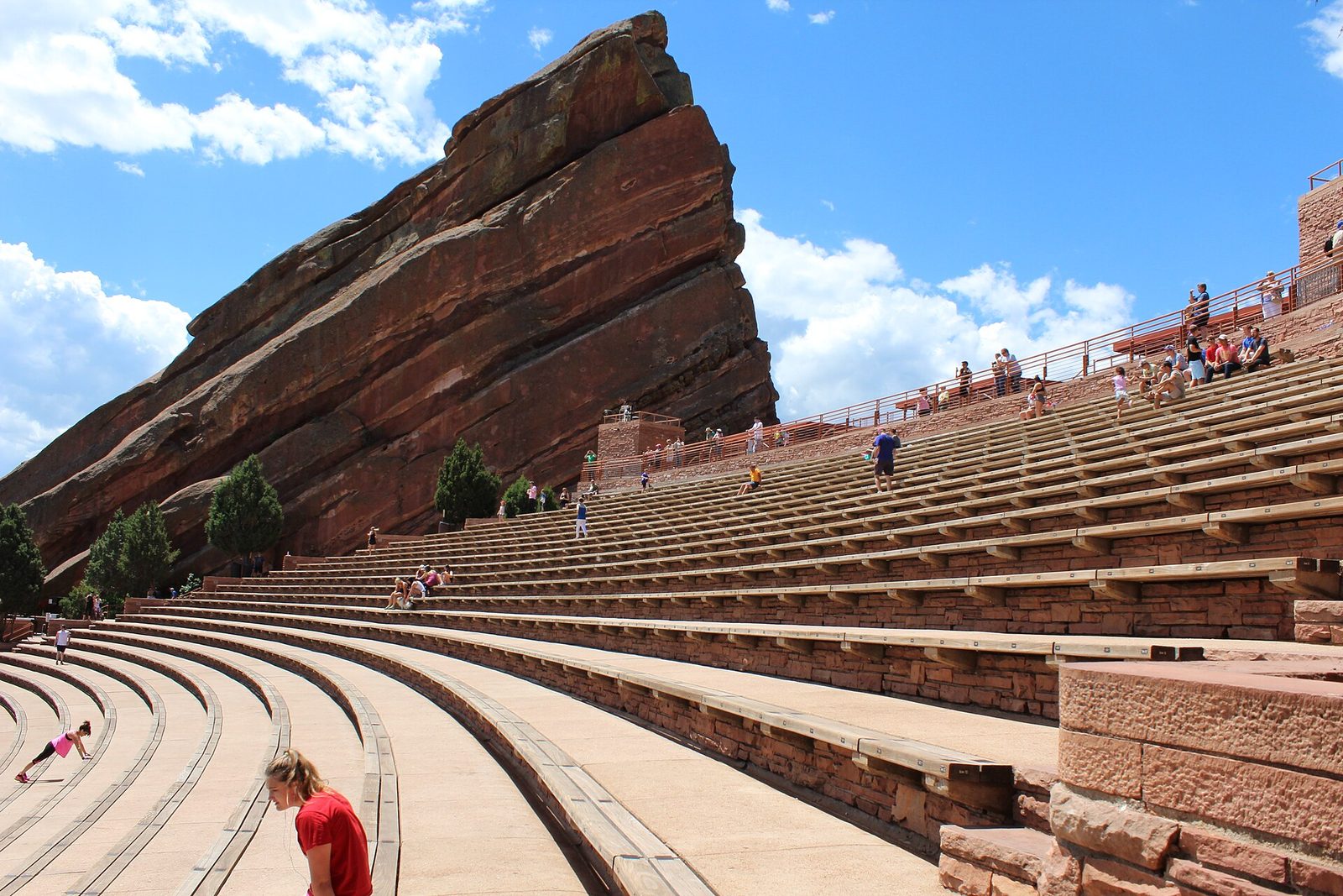 The tilted red sandstone slab of Creation Rock rising beside the curved bench seating of Red Rocks Amphitheatre, with people exercising on the steps