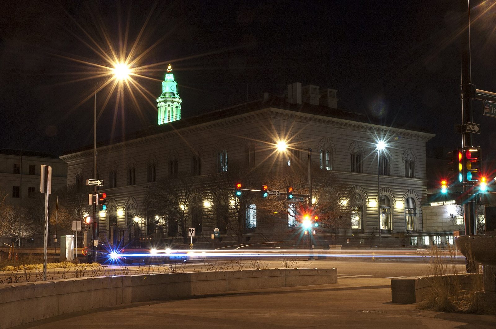 The stone Denver Mint building floodlit at night on a downtown corner, with arched ground-floor windows and the illuminated tower of the City and County Building rising behind it