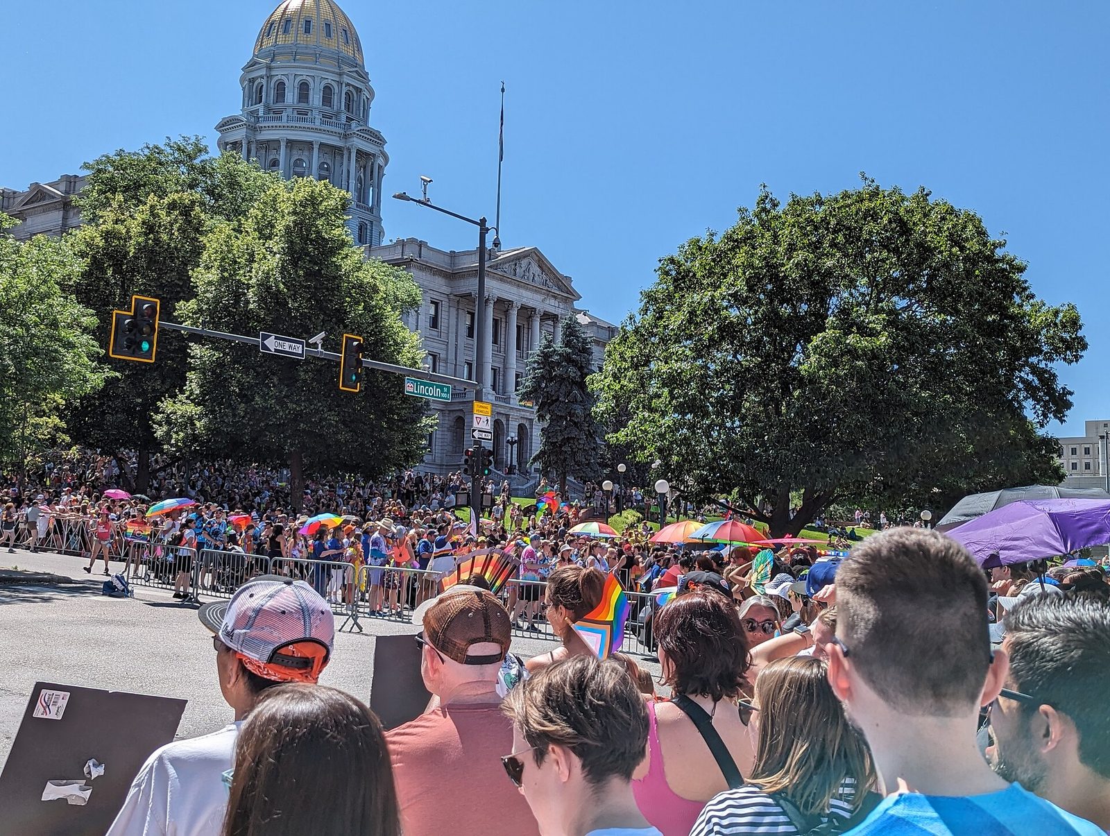 A dense crowd lining Lincoln Street in Denver for the PrideFest parade, with rainbow flags and umbrellas, and the gold dome of the Colorado State Capitol rising above the trees behind