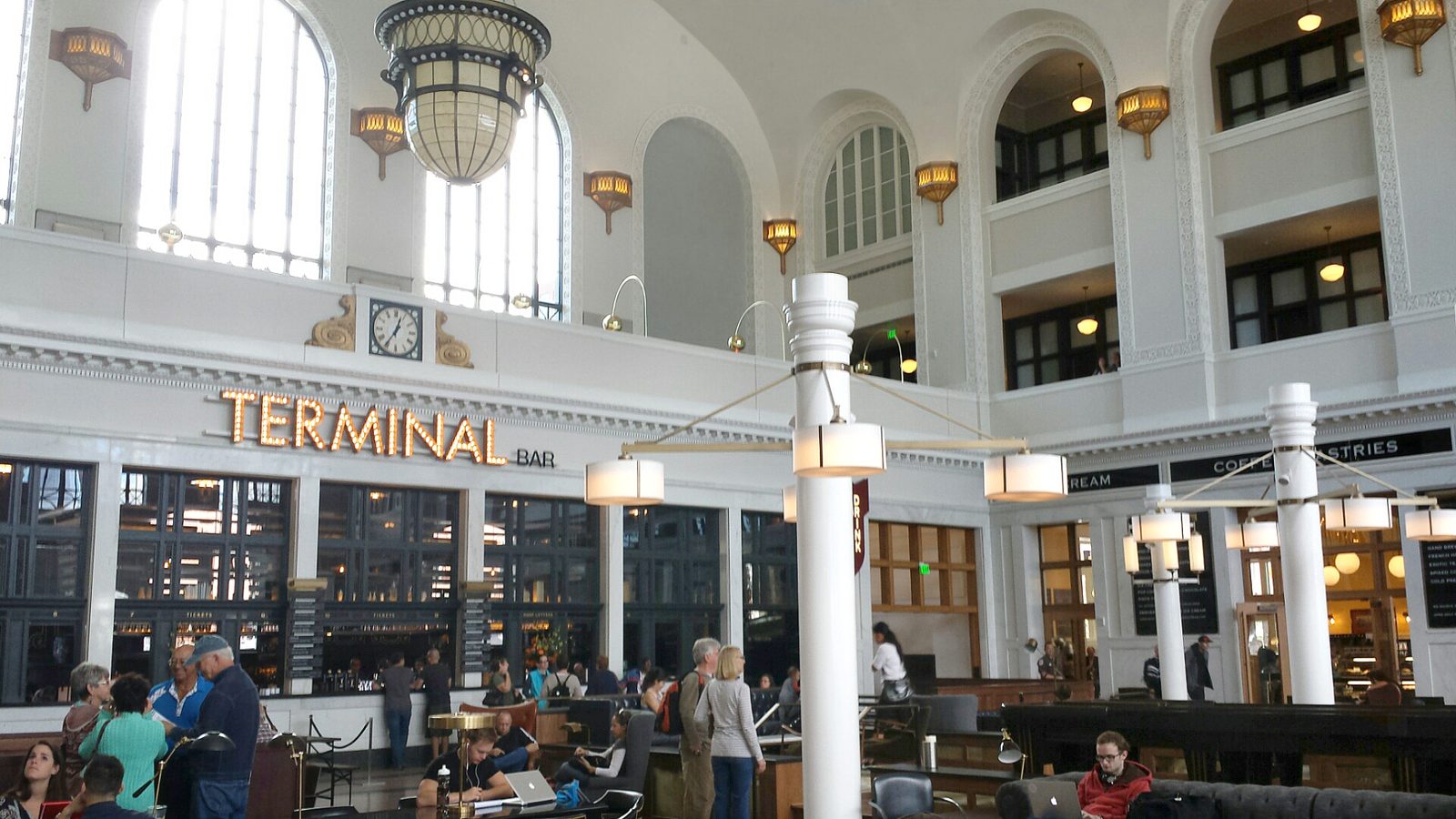 The Great Hall inside Denver Union Station, with tall arched windows, hanging lamps, a lit Terminal Bar sign and people sitting in leather armchairs at long tables