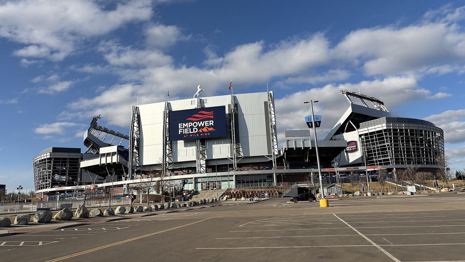 The exterior of Empower Field at Mile High in Denver on a clear winter day, with the stadium's video board and signage above an empty parking lot and the white bronco statue on the roofline