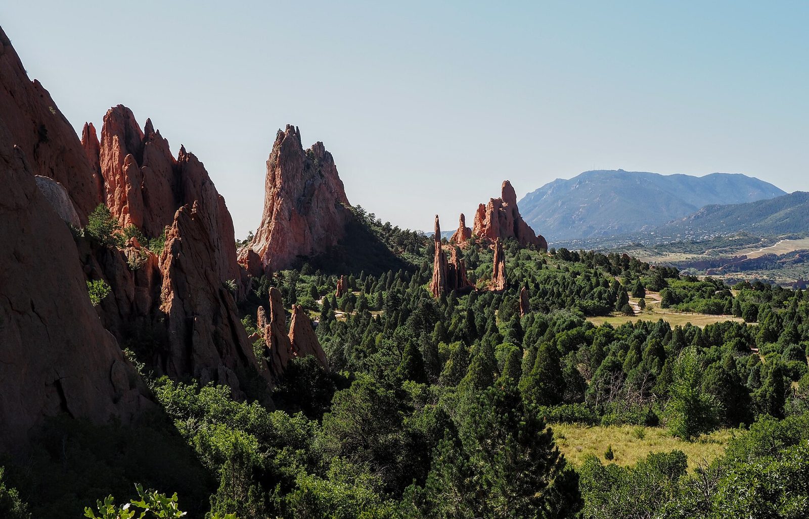 Red sandstone fins and spires rising out of green juniper scrub at Garden of the Gods in Colorado Springs, with a forested ridge and the city beyond