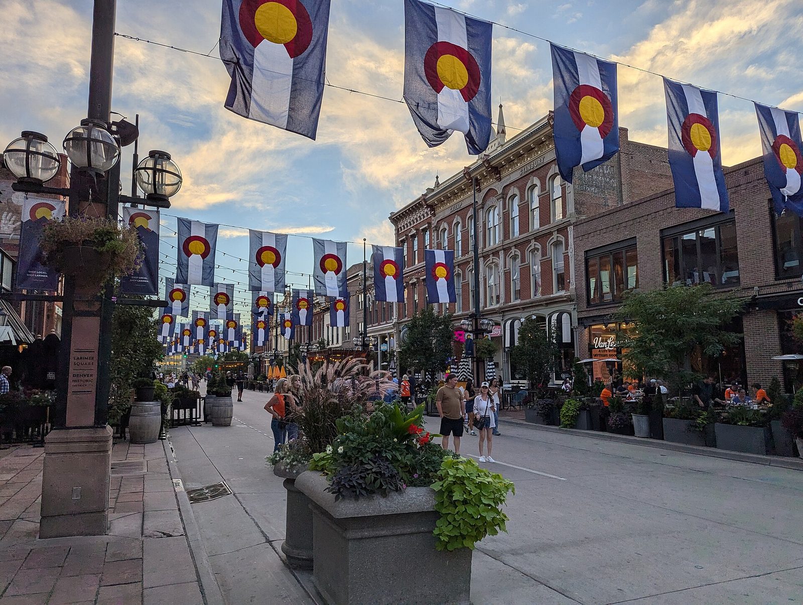 Colorado state flags and string lights hanging above the 1400 block of Larimer Street in Denver at dusk, with people walking and diners at patio tables