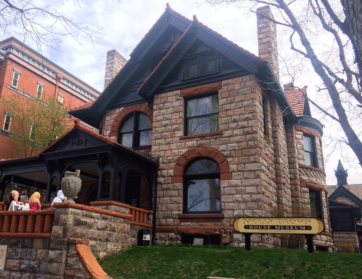 The stone and red sandstone Queen Anne exterior of the Molly Brown House Museum in Denver, with its museum sign on the lawn and visitors on the porch