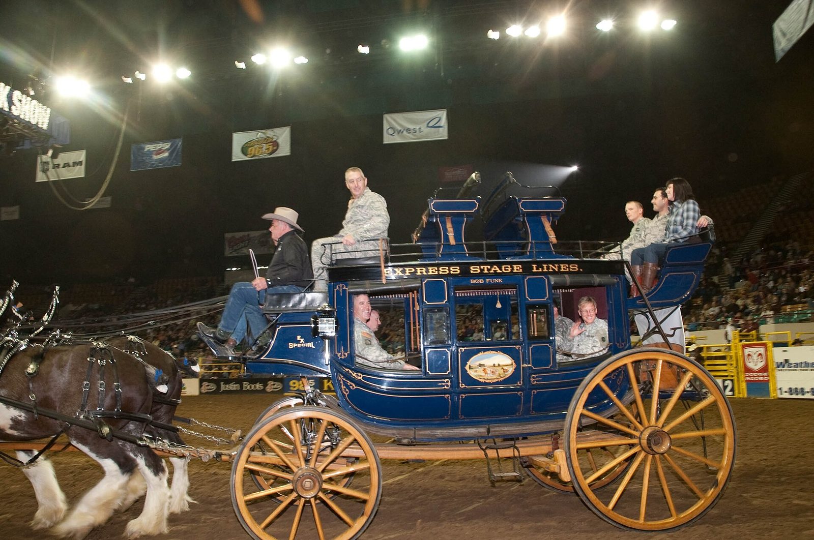 A team of draft horses pulling a restored blue nineteenth-century stagecoach with riders on top across the dirt arena floor at the National Western Stock Show in Denver, with spectators in the stands behind