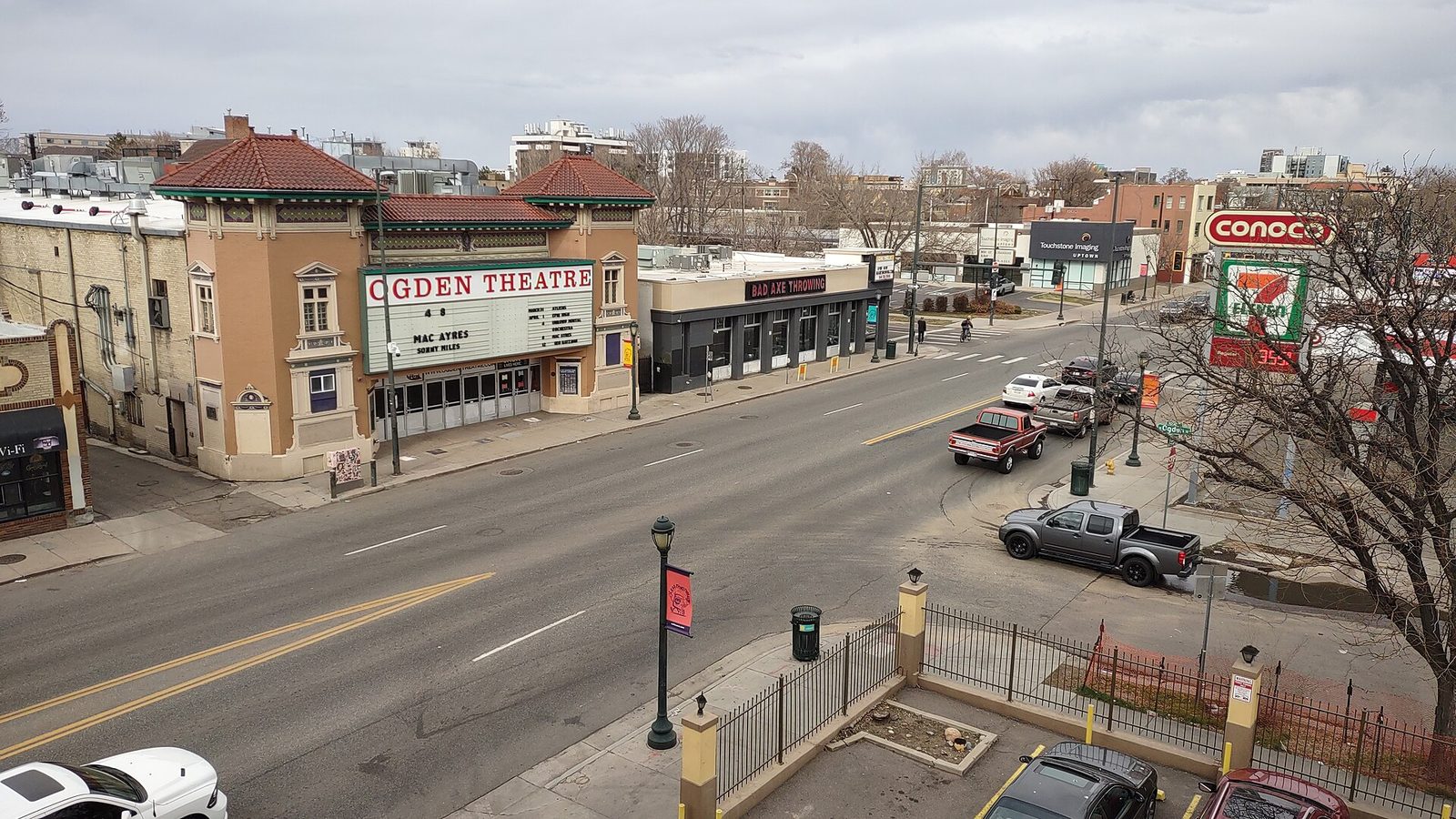 An elevated daytime view of East Colfax Avenue in Denver, with the Ogden Theatre and its marquee on the corner, low commercial buildings, a gas station and traffic on the wide street