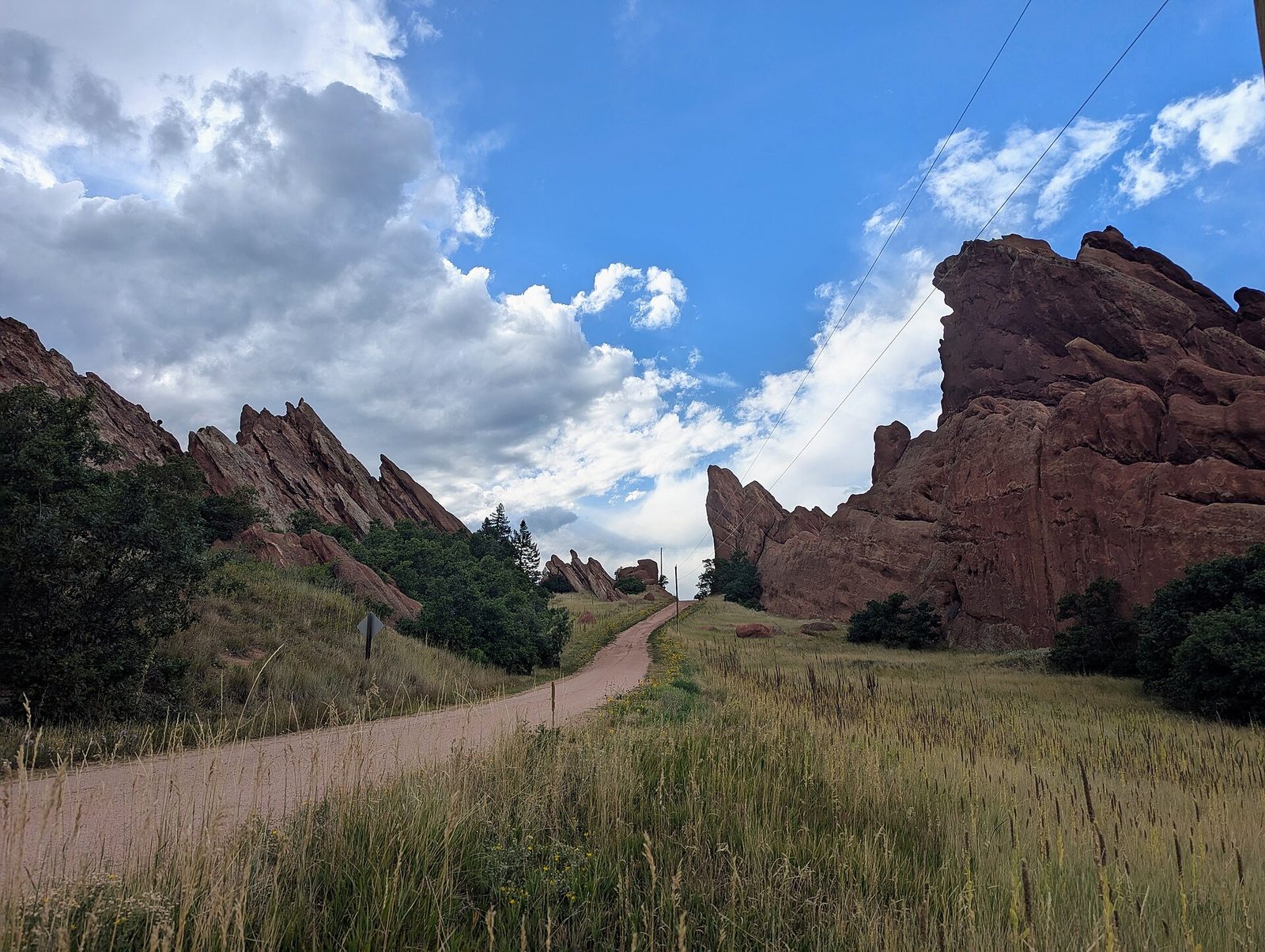 A gravel trail running between tilted red sandstone fins and summer grassland at Roxborough State Park south of Denver, under a sky of building cumulus