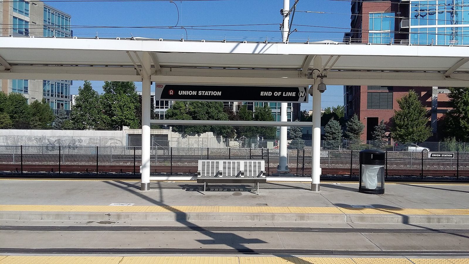 An RTD light rail platform at Union Station in Denver with an overhead sign reading Union Station, End of Line, a bench under the canopy and apartment buildings behind