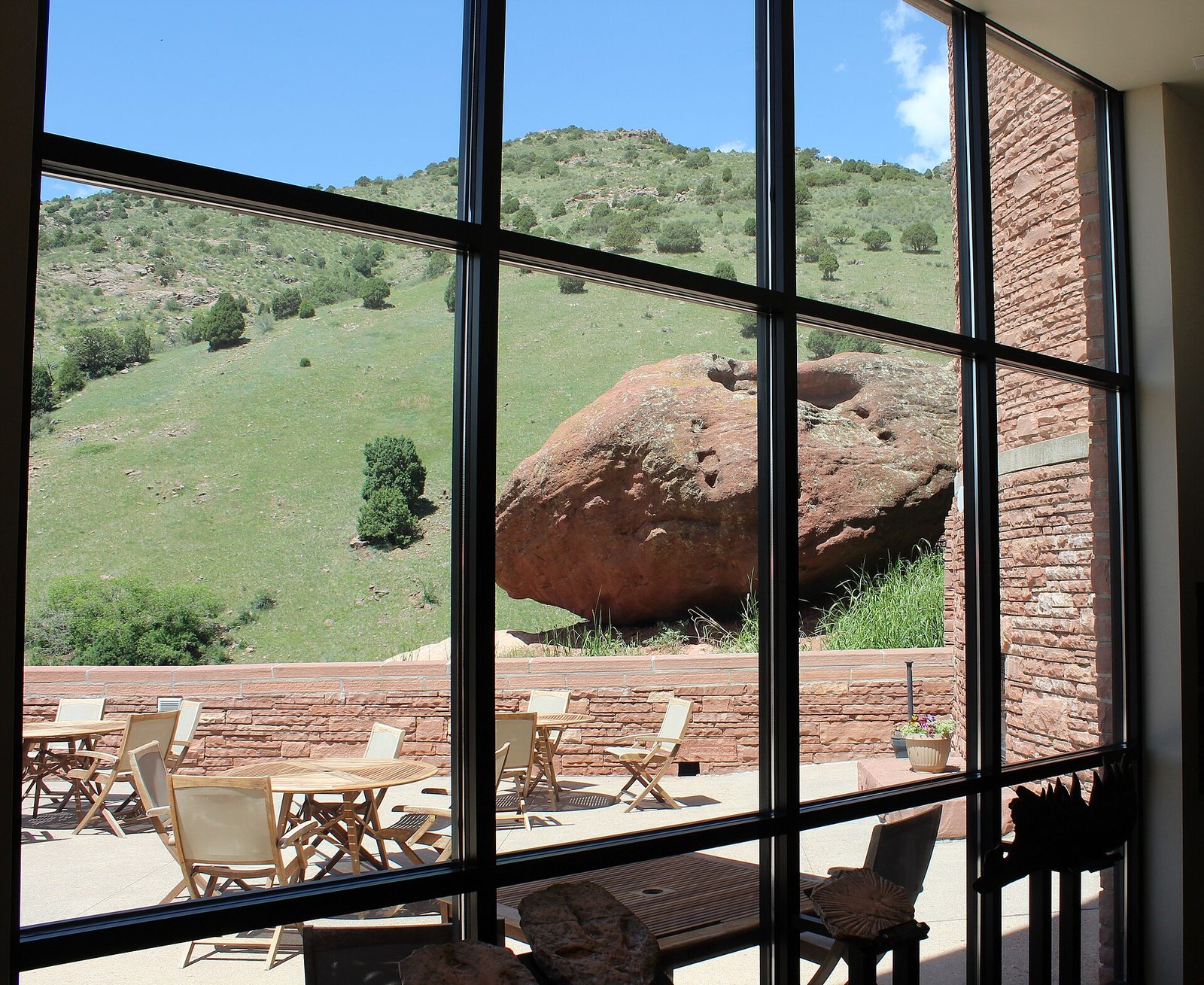 The interior of the Ship Rock Grille at Red Rocks, looking out through a full-height glass wall at a red sandstone boulder and a green hillside, with patio tables outside