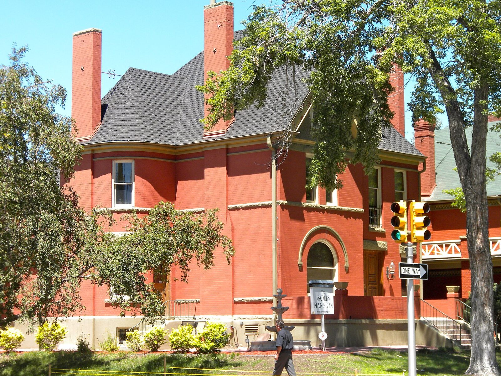 A large red brick Victorian mansion with tall chimneys on a corner in the Swallow Hill historic district of Denver, with mature trees around it