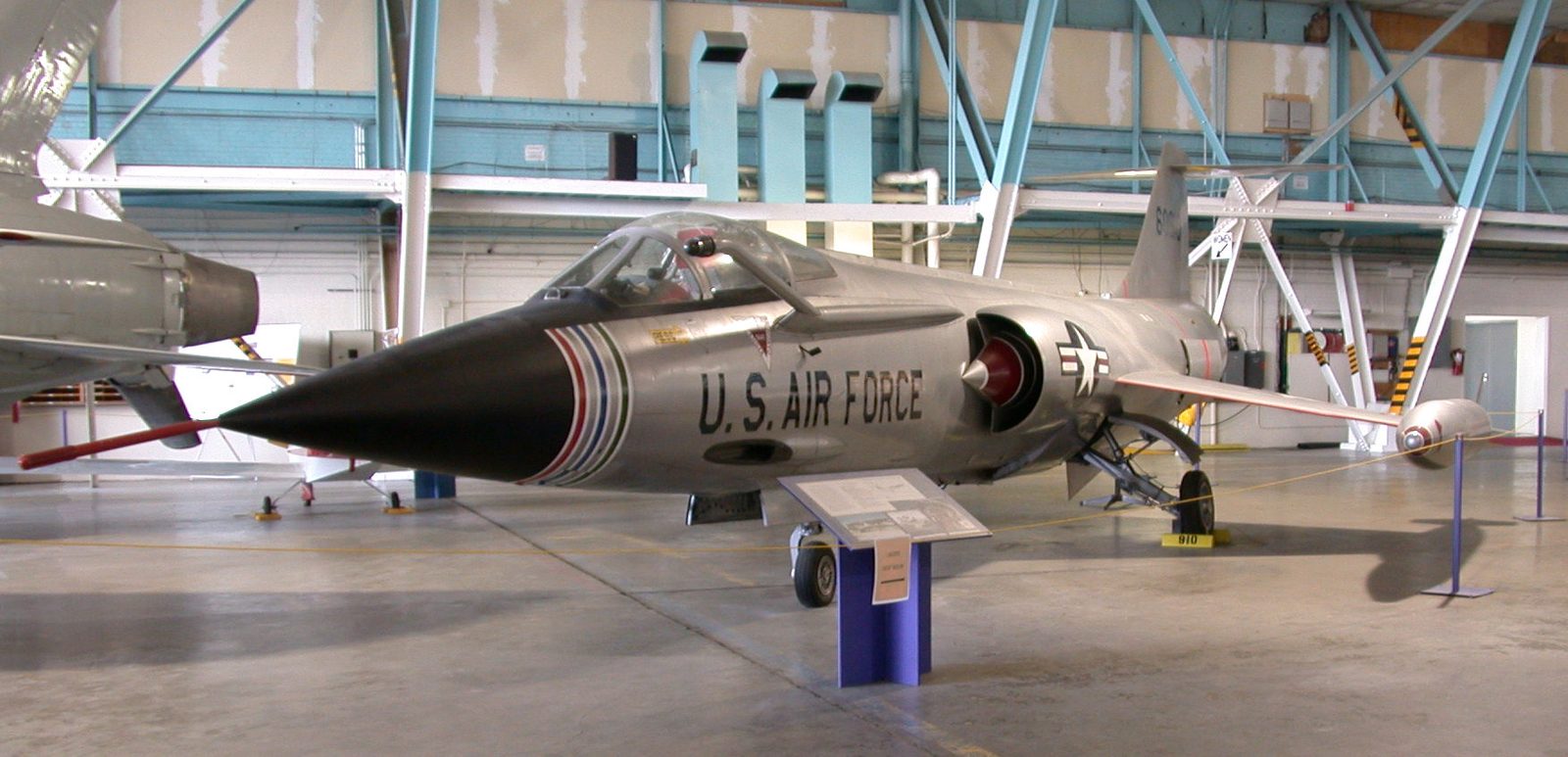 A silver Lockheed F-104C Starfighter jet on display inside the Wings Over the Rockies museum hangar in Denver, with other aircraft behind it and an information panel in front