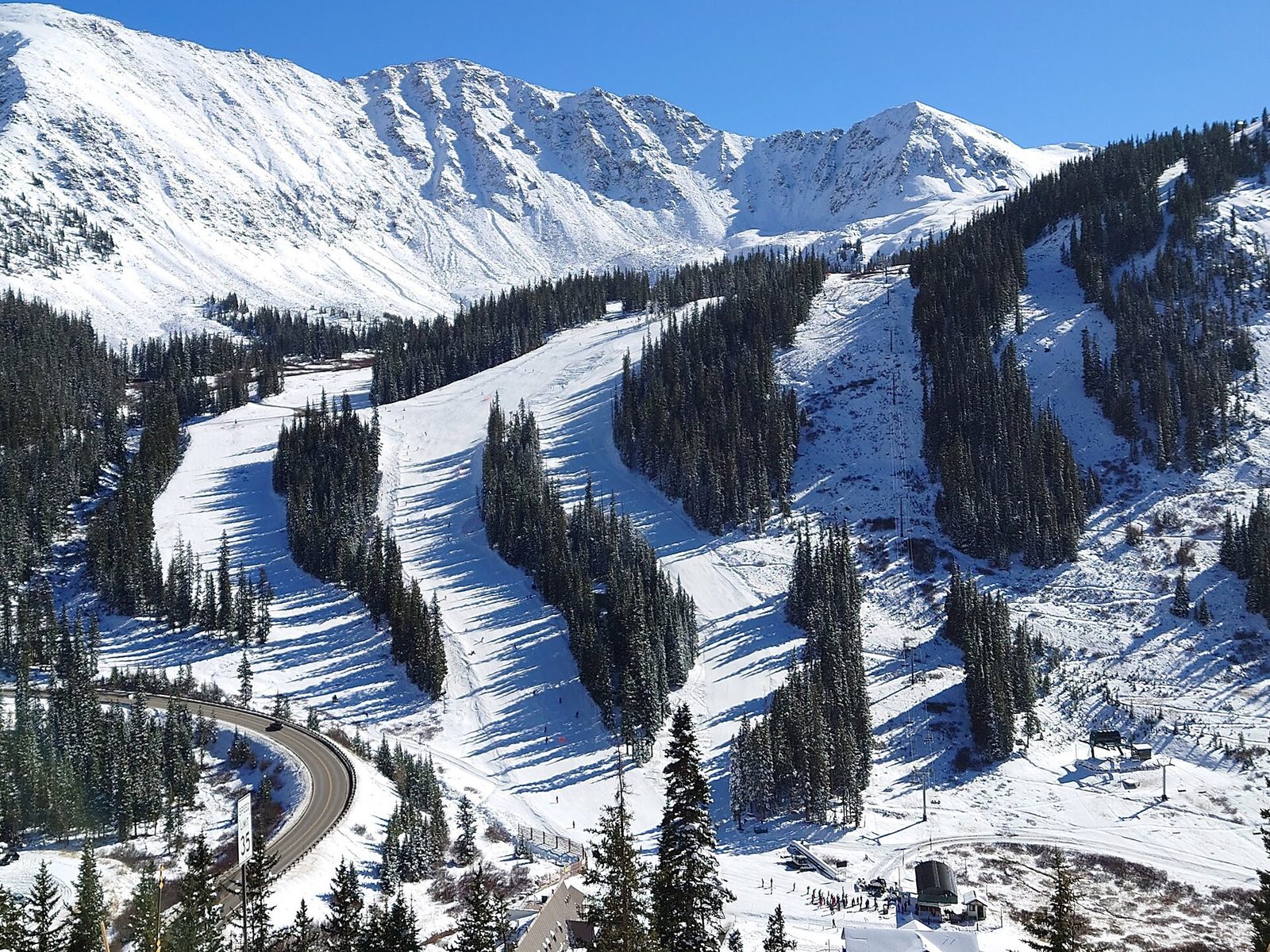 The base area and lower trails of Arapahoe Basin a few days after opening in late October, with white ribbons of snow cut through dark conifers, a steep snowy ridge above and the curve of US 6 in the foreground
