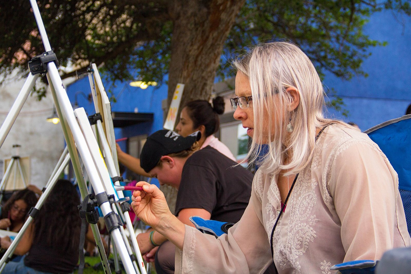 A woman with long fair hair and glasses painting on a canvas at an easel outdoors on Santa Fe Drive in Denver, with other painters at easels behind her under a tree in front of a blue-painted building