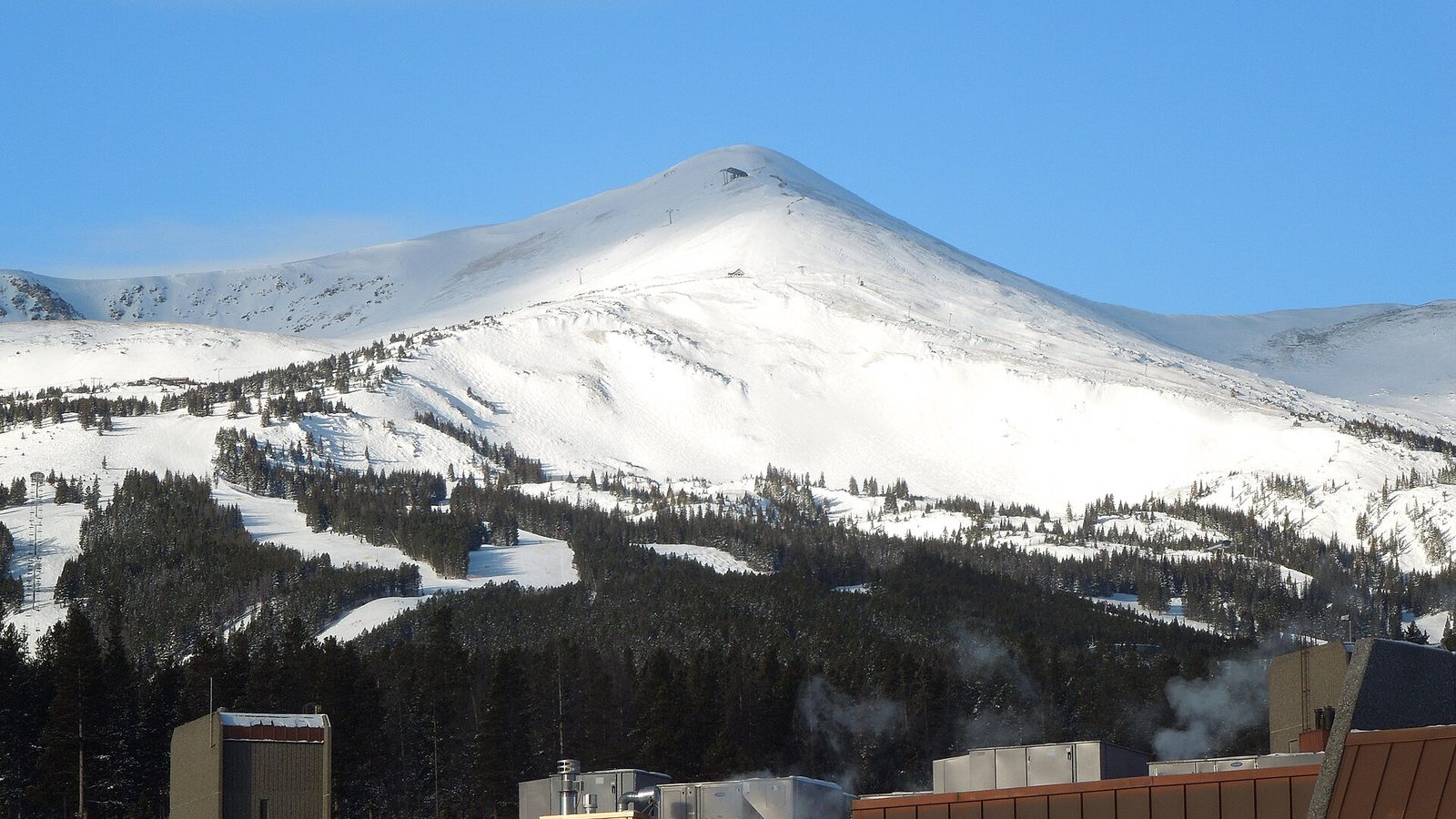 The snow-covered summit of Peak 8 at Breckenridge Ski Resort under a clear blue sky, with ski runs cut through the conifers on its lower slopes and rooftops of the base area in the foreground