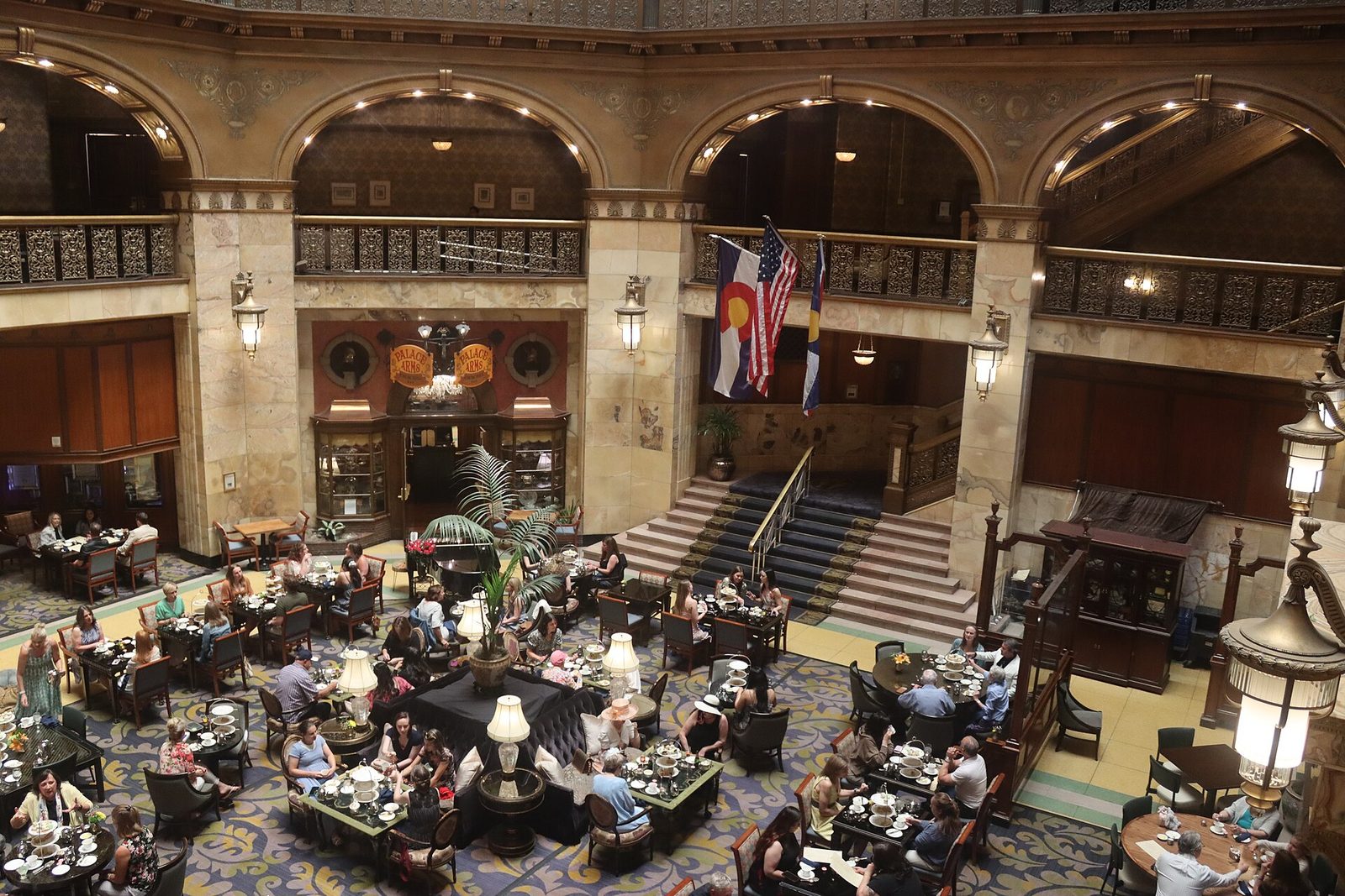 The atrium lobby of the Brown Palace Hotel in Denver seen from a balcony, with guests at tables set for afternoon tea, palms, lamps, a carpeted staircase and ornate iron balconies under tall arches