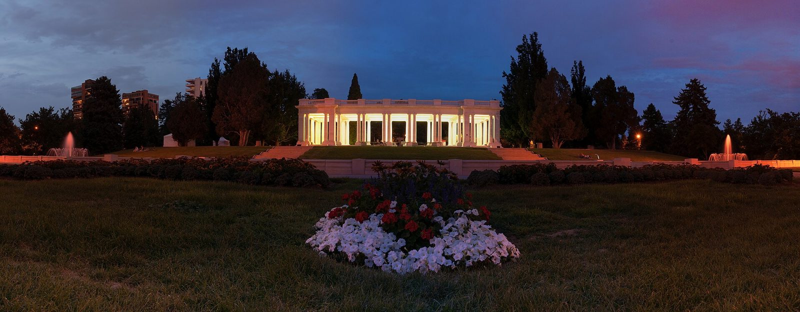 The white marble colonnaded pavilion in Cheesman Park in Denver lit up at dusk, with fountains on both sides, dark trees and apartment towers behind, and a bed of red and white flowers on the lawn in front