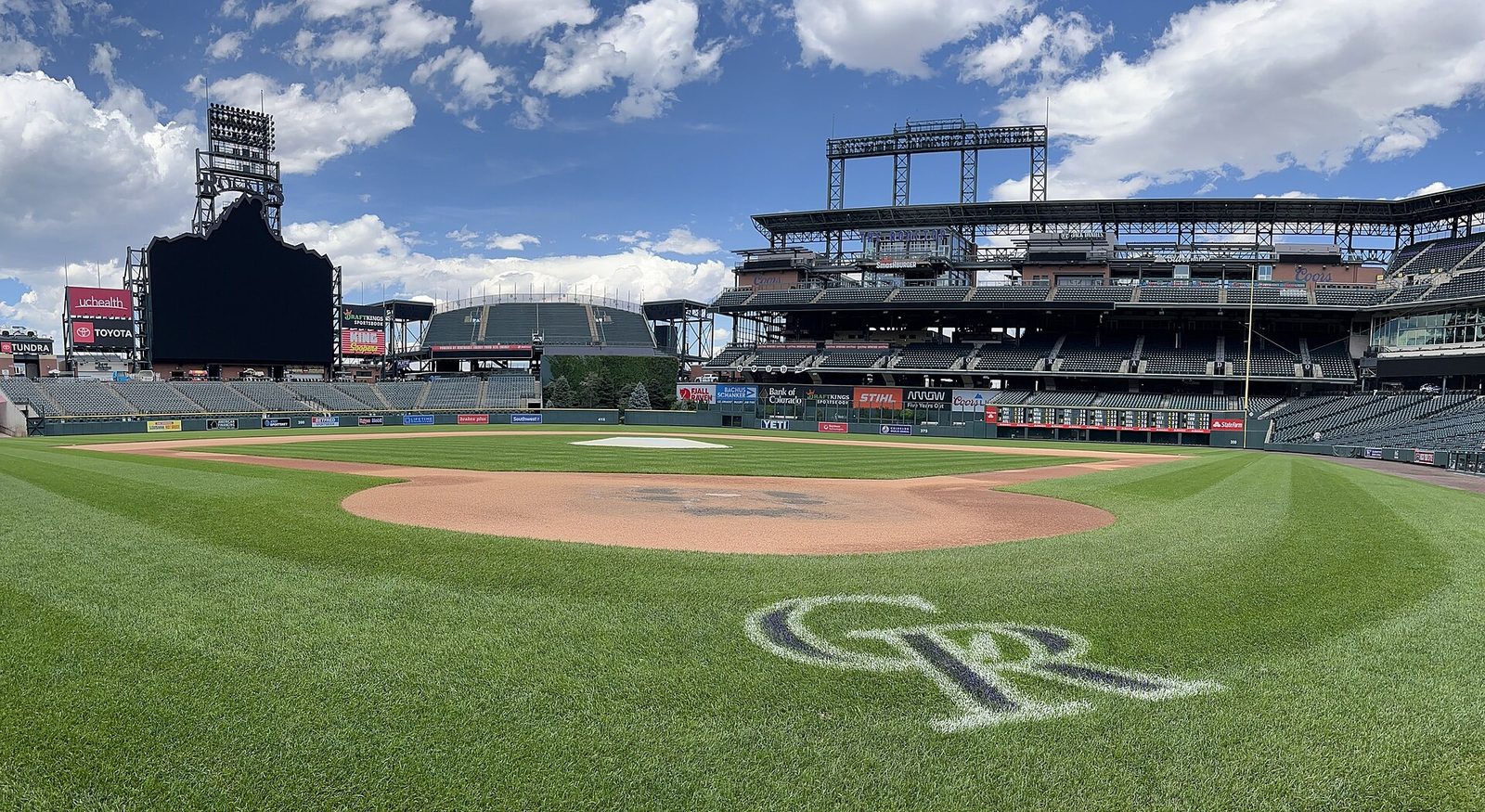 A wide view of Coors Field from behind home plate on a sunny day, with the Rockies logo mown into the grass, the empty green seating bowl and the scoreboard under white clouds