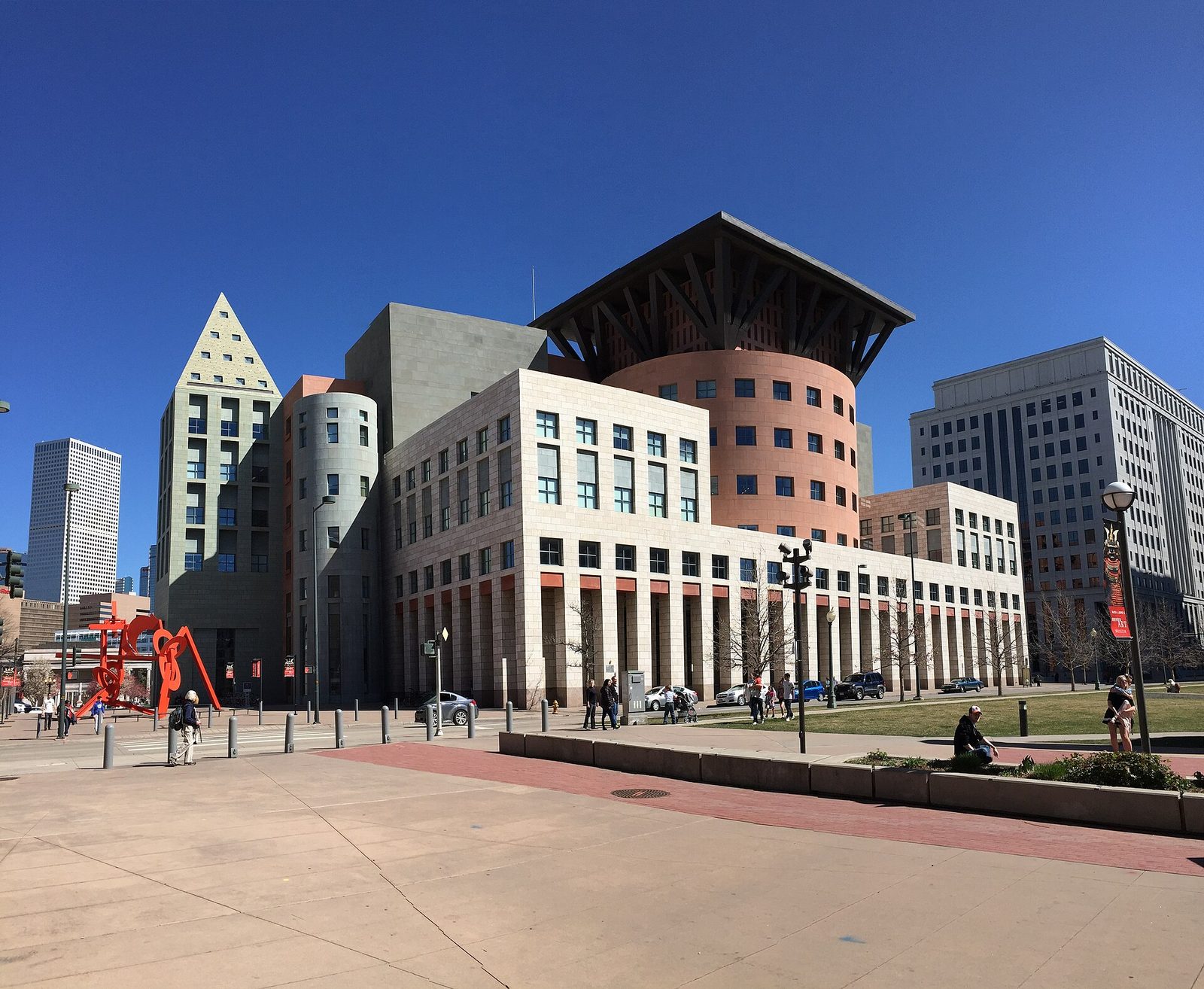 The Central Library of the Denver Public Library designed by Michael Graves, a cluster of pastel-colored towers and a copper-roofed drum, with a large red steel sculpture on the plaza to the left and downtown towers behind under a deep blue sky
