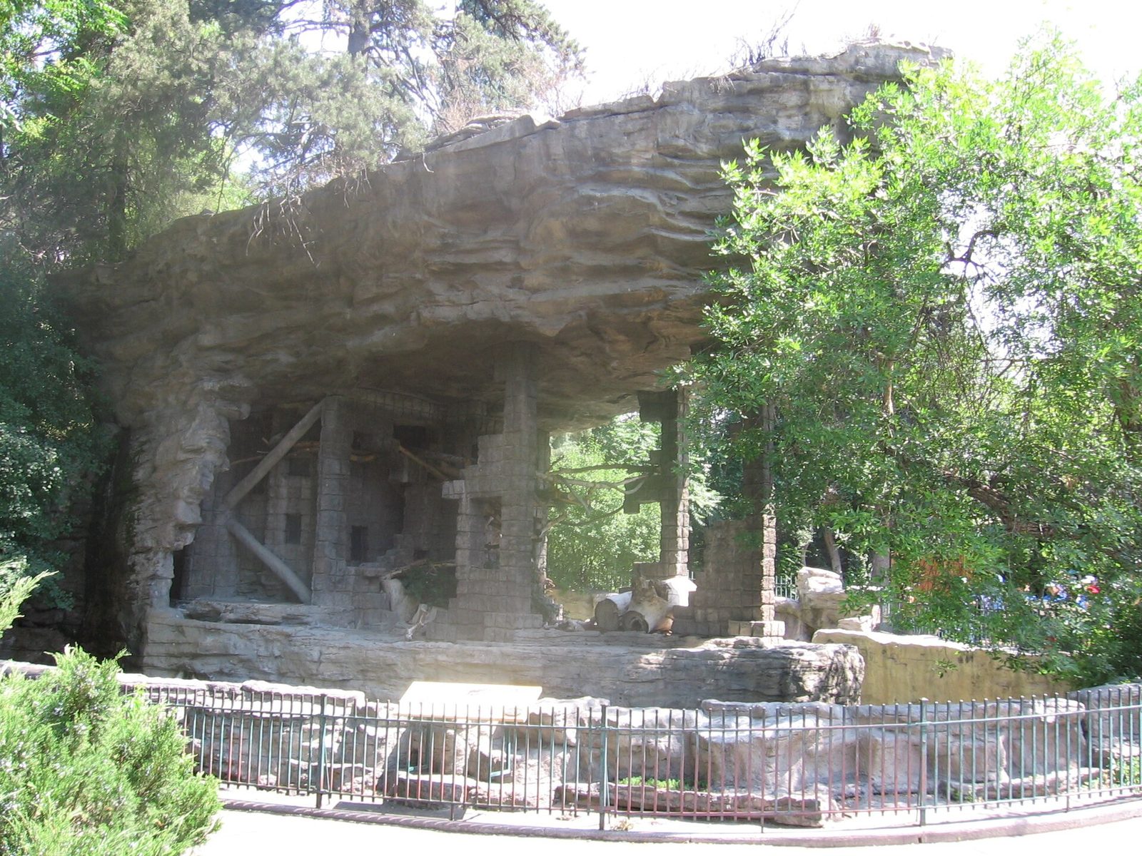 Bear Mountain at the Denver Zoo, a large overhanging cliff of sculpted concrete made to look like natural rock, with stone pillars, logs and an iron railing in front, framed by trees