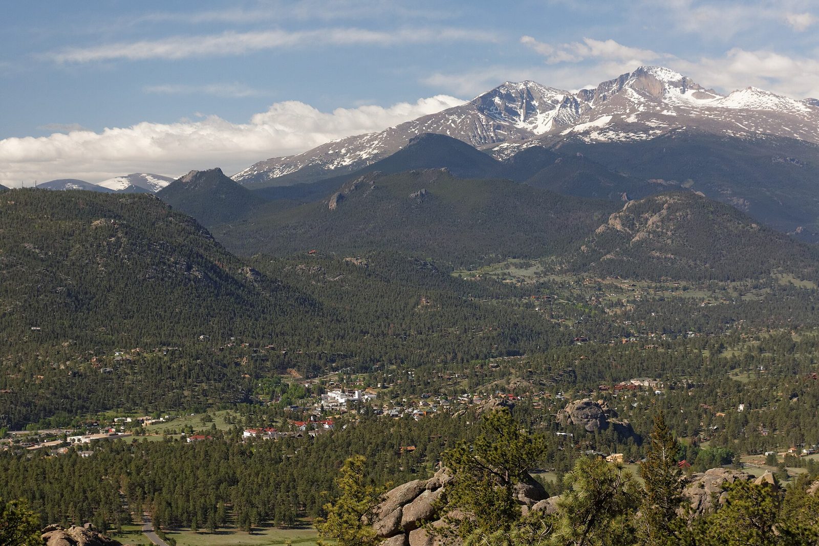 The town of Estes Park spread across a forested valley below snow-streaked Longs Peak and Mount Meeker, seen from a rocky viewpoint on a sunny late-spring day