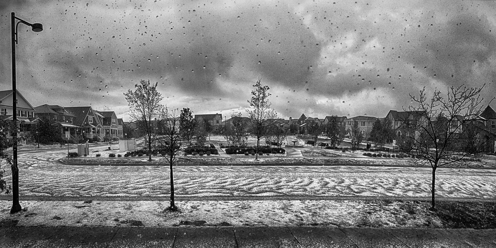 A black and white photograph of a park in a Denver neighborhood covered in drifts of hail during a sudden storm, with houses around the green, bare young trees, a streetlamp and hailstones streaking through the air