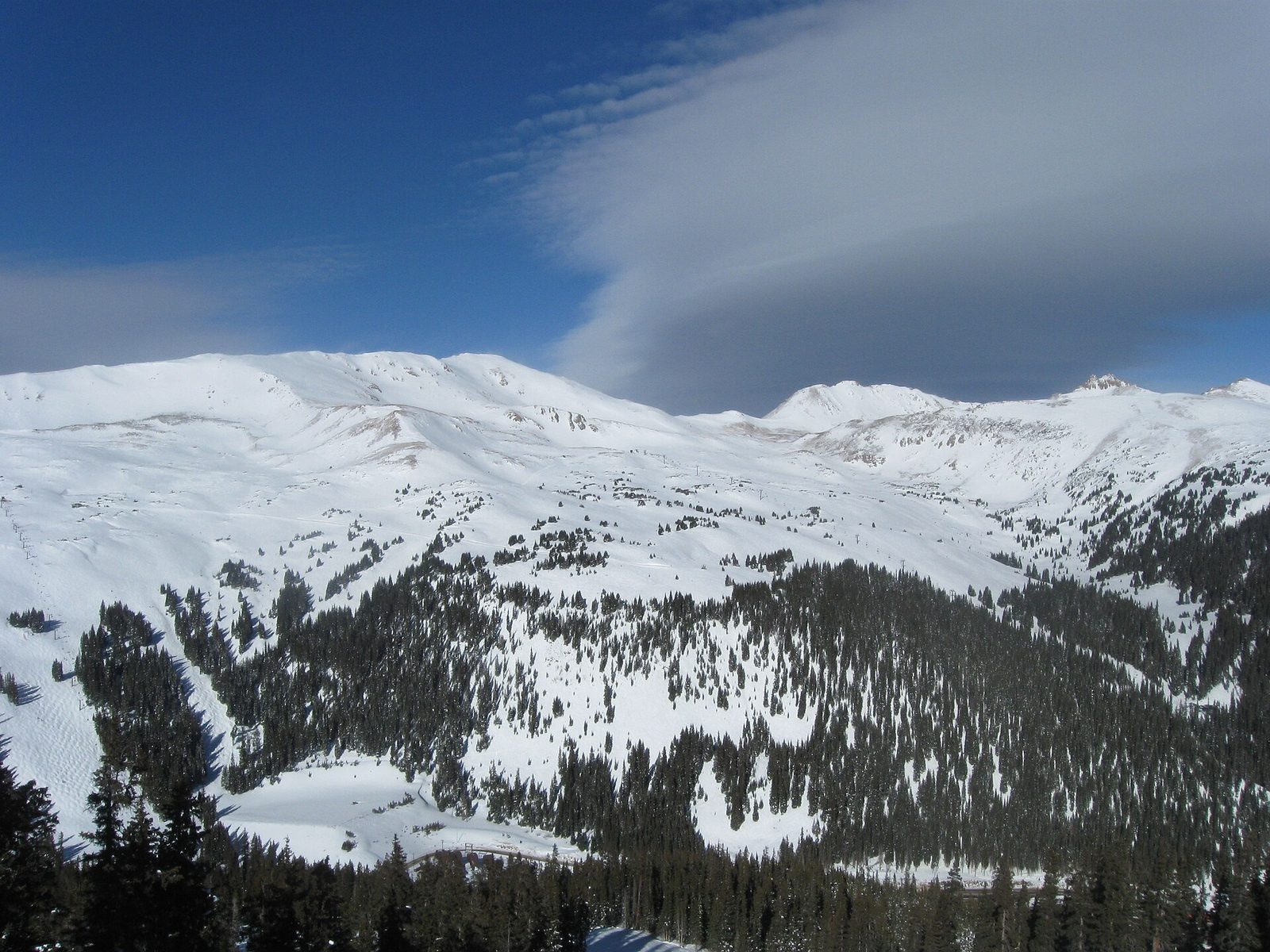 Snow-covered bowls and ridges above the tree line at Loveland Ski Area on the Continental Divide, with scattered conifers on the lower slopes and a long lens-shaped cloud over a deep blue sky