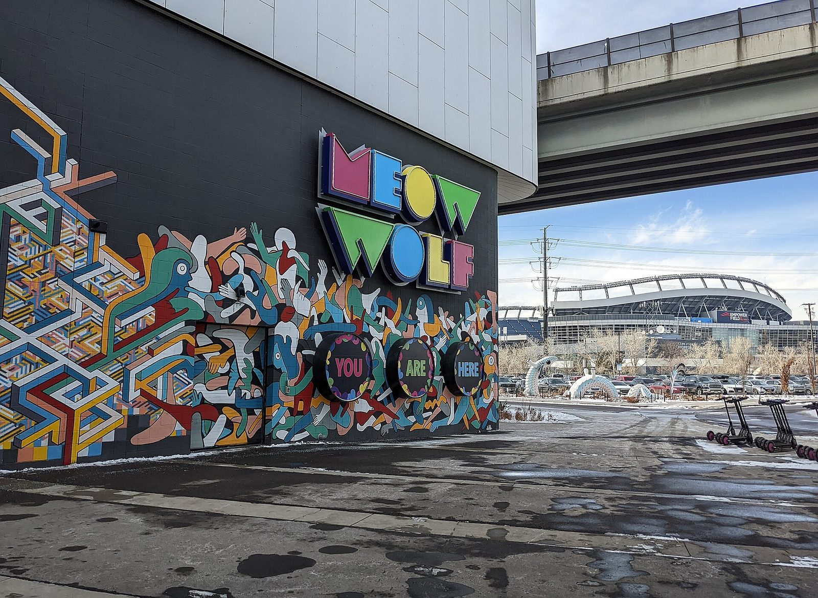 The colorful Meow Wolf sign and a large mural reading You Are Here on the wall of Convergence Station in Denver, beneath an I-25 overpass, with Empower Field at Mile High in the background on a winter day