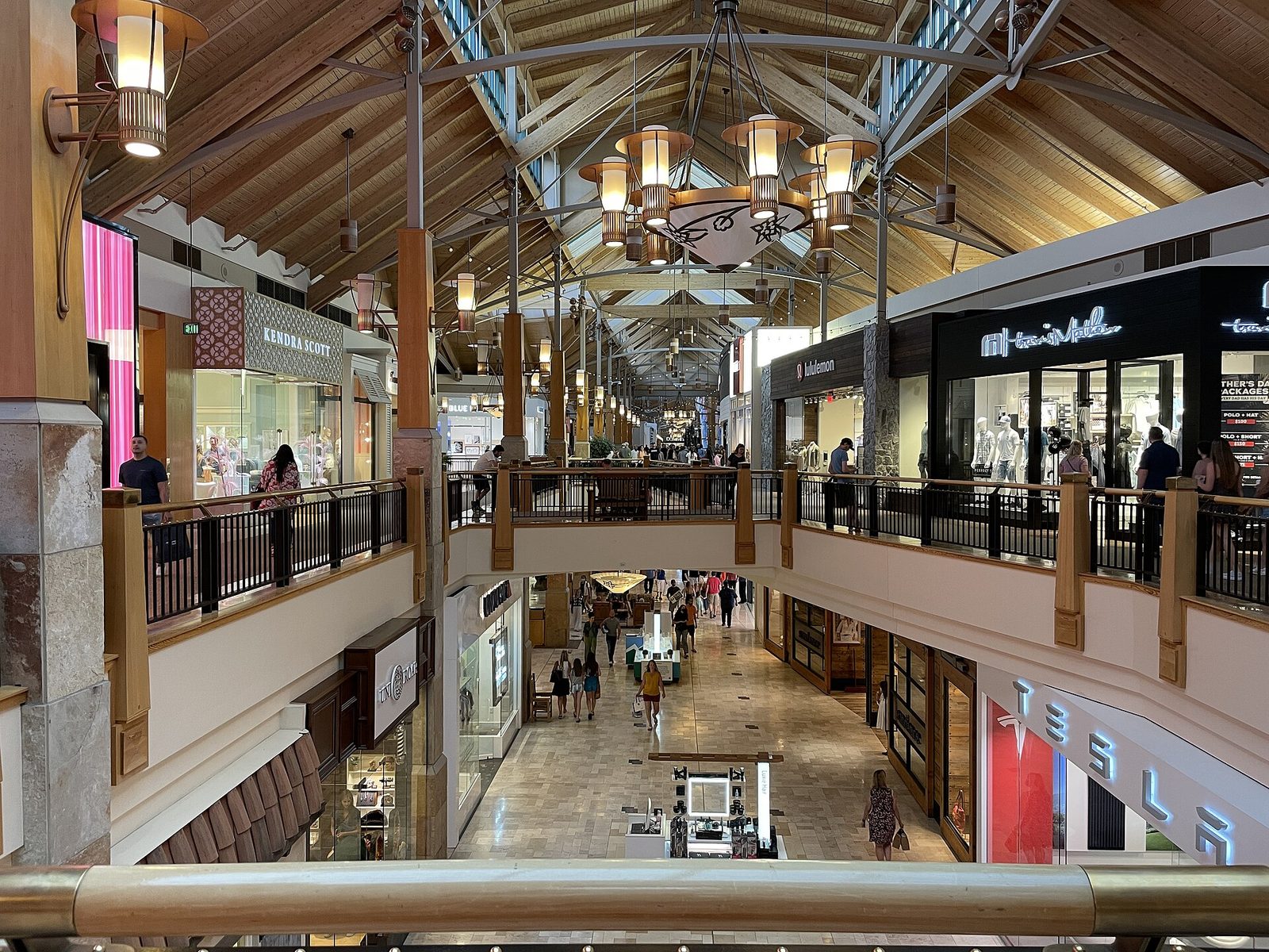 The two-level interior of Park Meadows mall in Lone Tree, Colorado, with a pitched timber ceiling, skylights, lantern chandeliers, wooden railings and shoppers walking past storefronts below