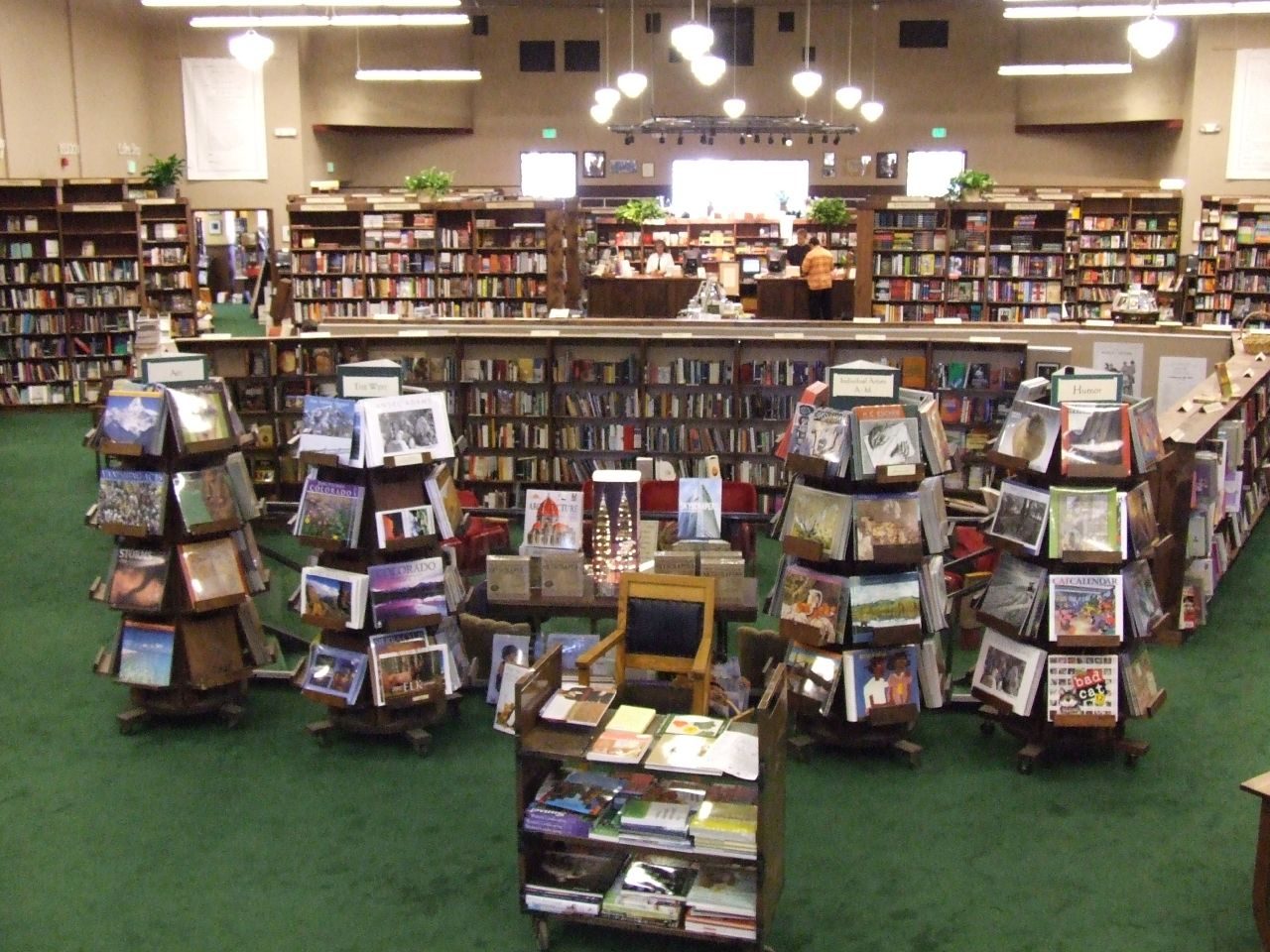 The main floor of the Tattered Cover bookstore on East Colfax Avenue in Denver, with green carpet, rotating display stands of calendars and photography books in front, wooden shelves behind and staff at a central counter under hanging lamps