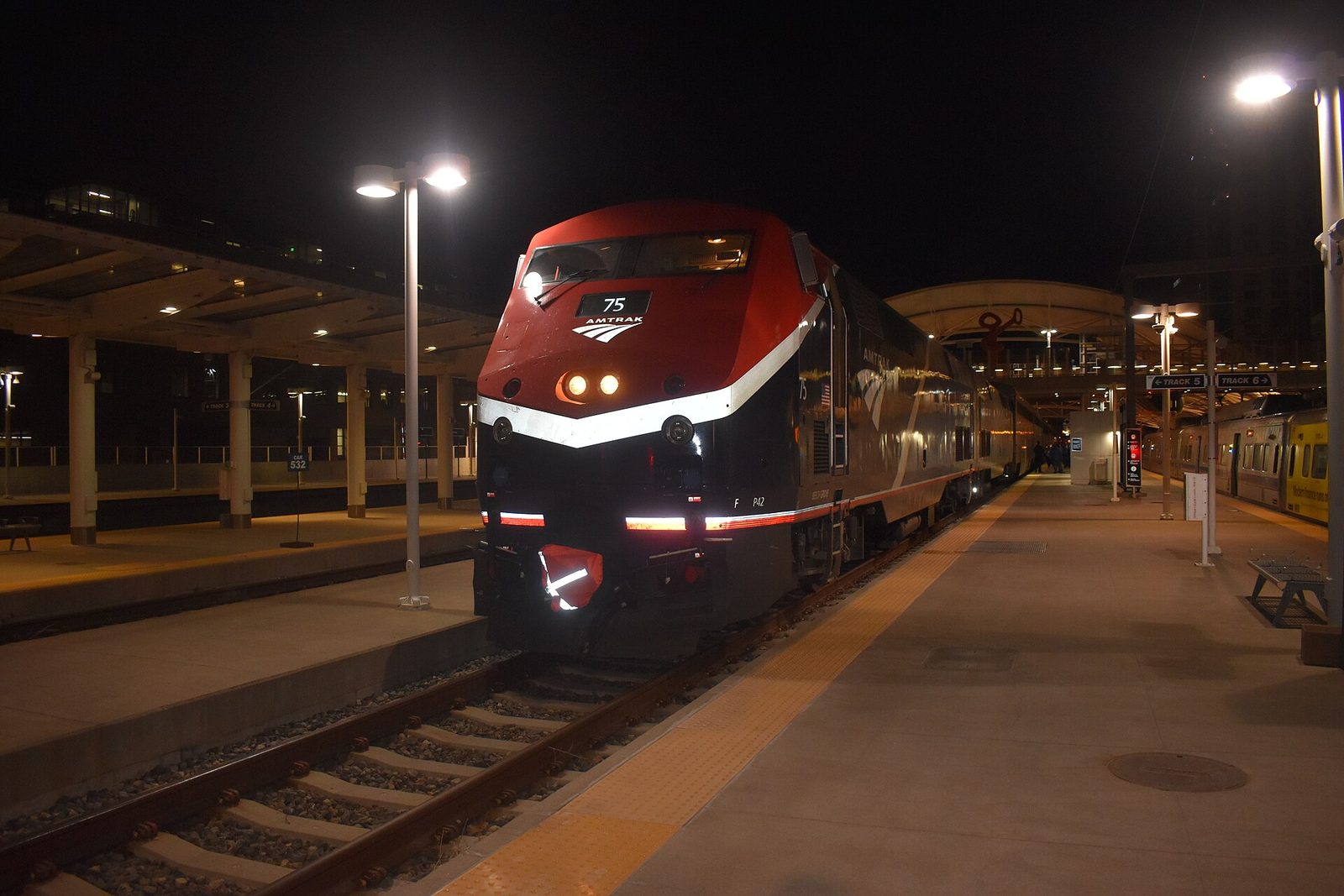 An Amtrak locomotive numbered 75 at the head of the Winter Park Express at a lit platform at Denver Union Station before dawn, with the station canopy and a commuter train on the next track